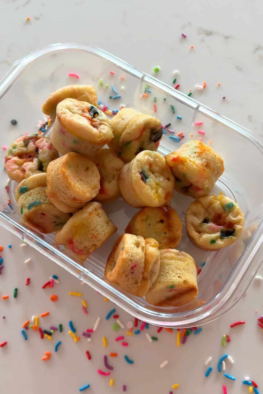 Greek yogurt sprinkle mini muffins stored in a container with rainbow sprinkles scattered around it
